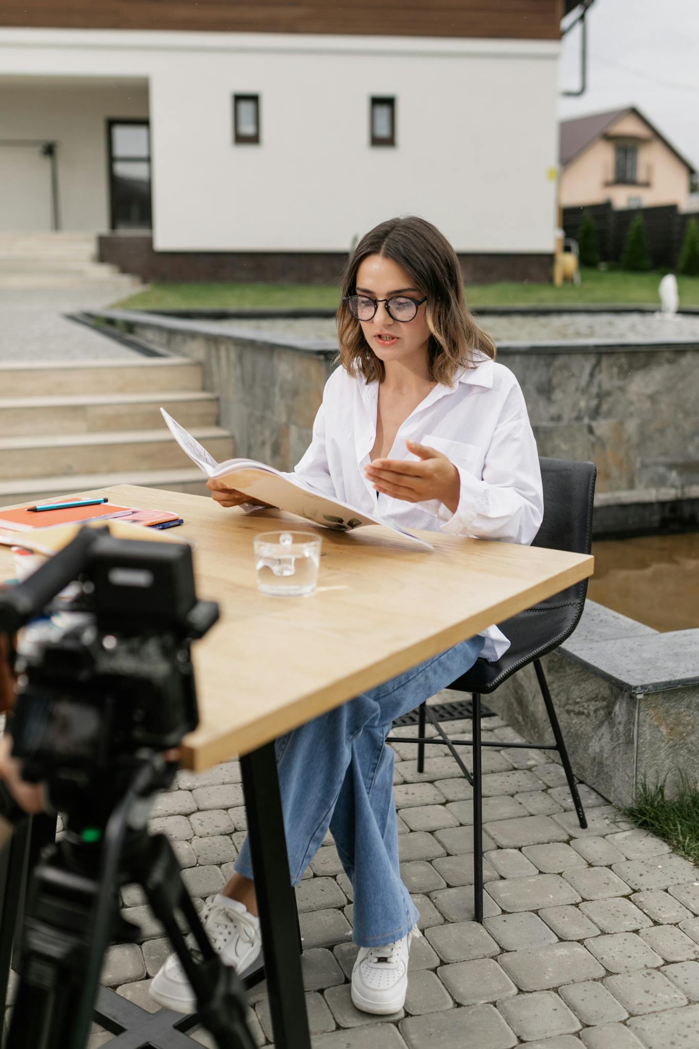 A woman in eyeglasses sitting outdoors while reading and vlogging at a table.